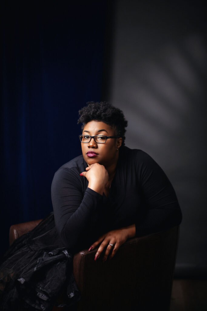 Professional portrait of author Vera T. Brown, seated and leaning on her hand, wearing glasses and a black top.